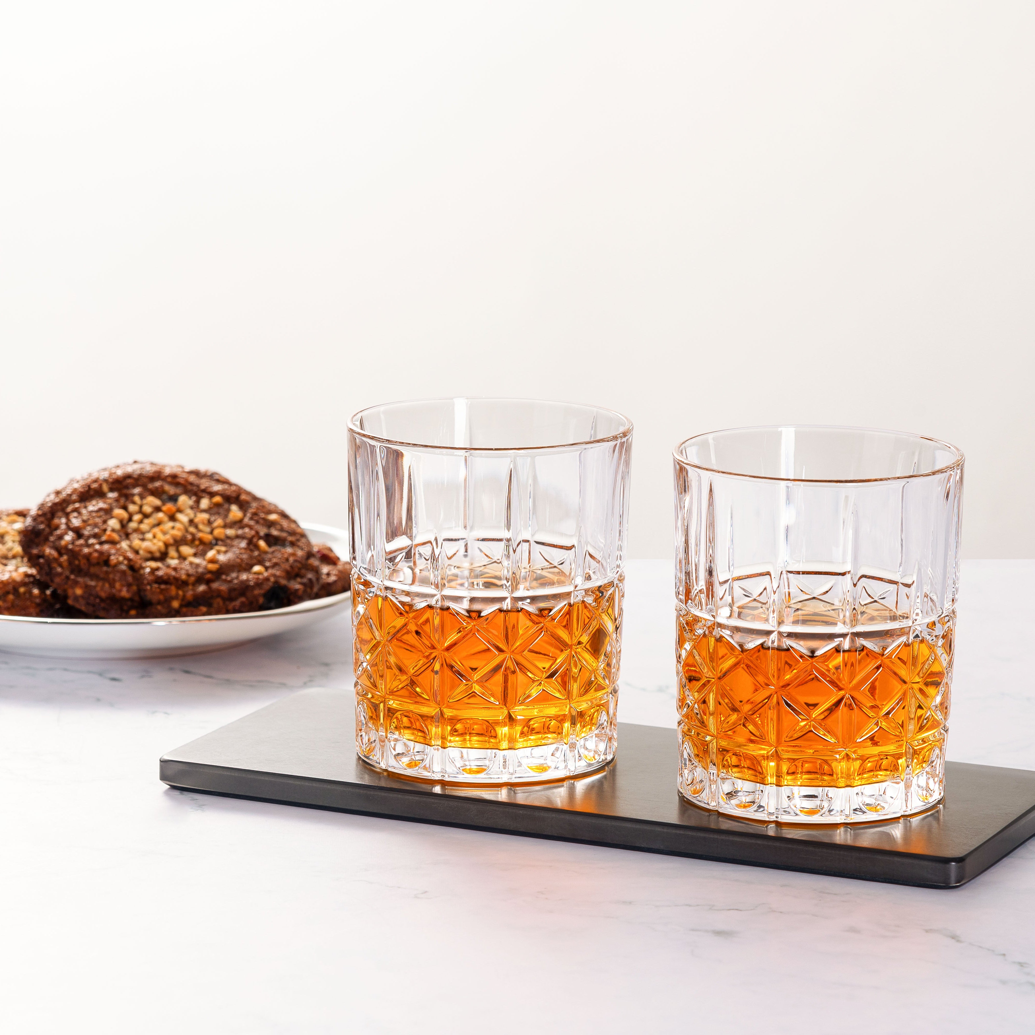 Load image into Gallery viewer, Two glasses of whisky on a black coaster with cookies on a white plate in the background.
