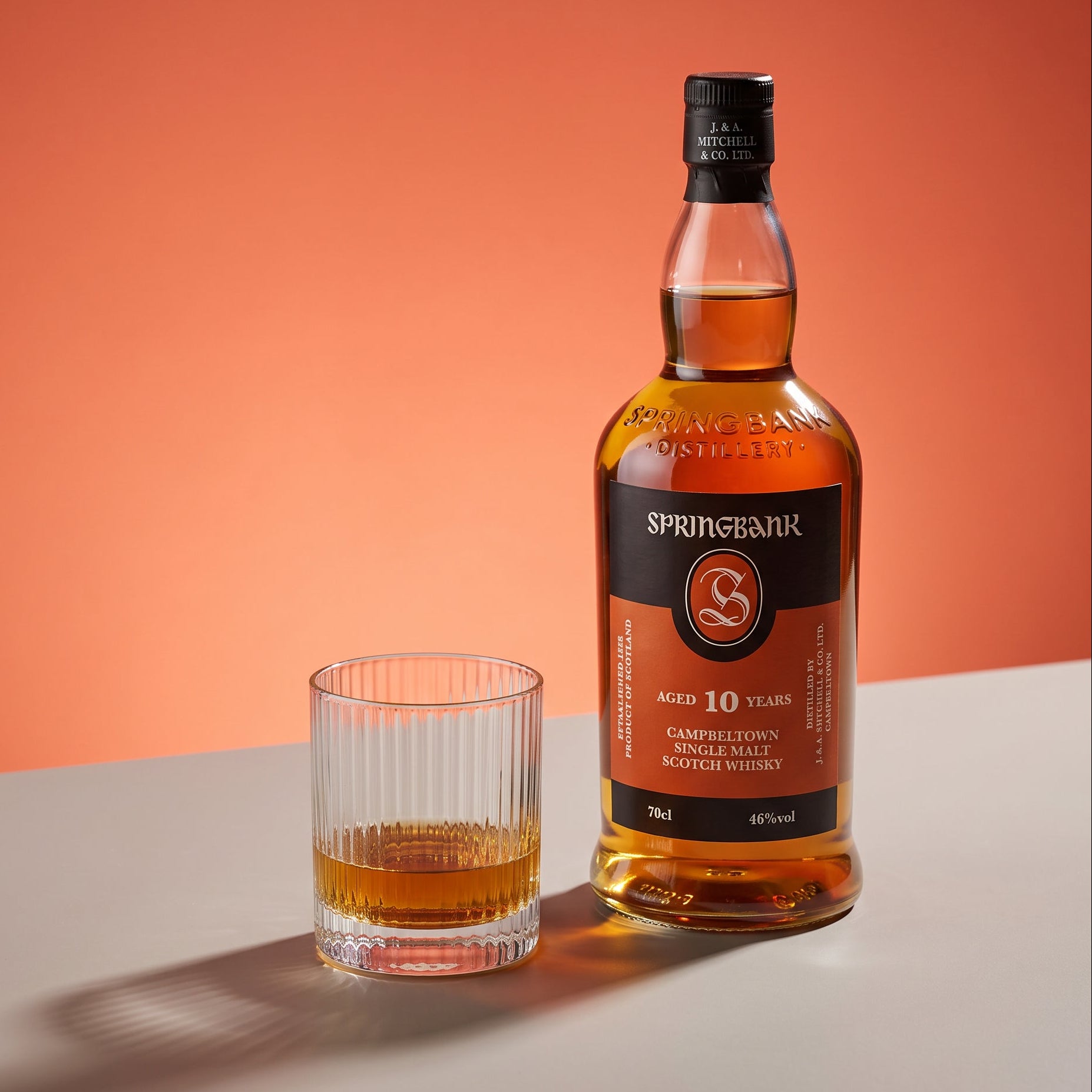 Load image into Gallery viewer, A bottle of Springbank 10 Years Old single malt Scotch whisky stands on a white surface next to a clear glass tumbler containing some of the whisky, against a solid orange background.