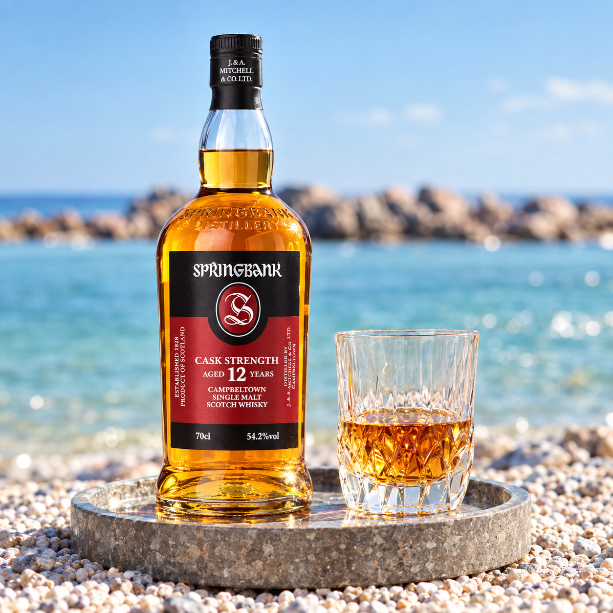 Load image into Gallery viewer, A bottle of Springbank Cask Strength 12 Year Old Scotch Whisky stands on a stone tray beside a filled coupe glass, set on a pebble beach with the blue sea and sky blurred in the background.