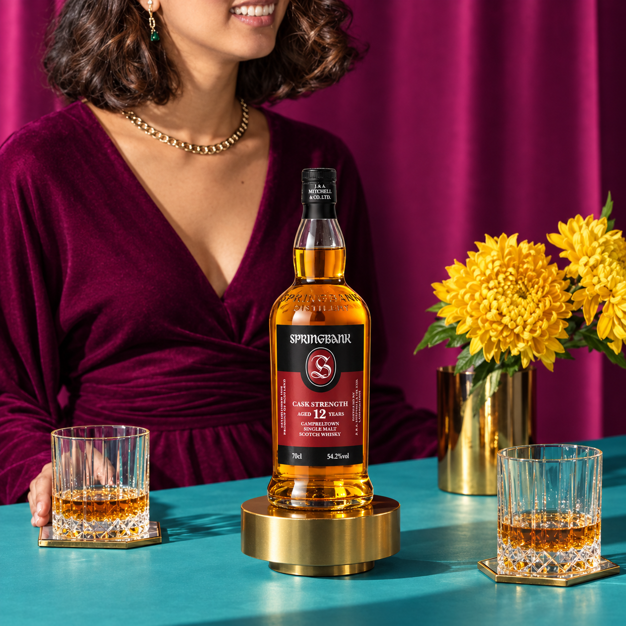 Load image into Gallery viewer, A bottle of Springbank Cask Strength 12 Year Old Single Malt Scotch whisky is displayed on a gold pedestal on a teal table, with two glasses of whisky, yellow flowers, and a woman in a purple velvet dress in the background.