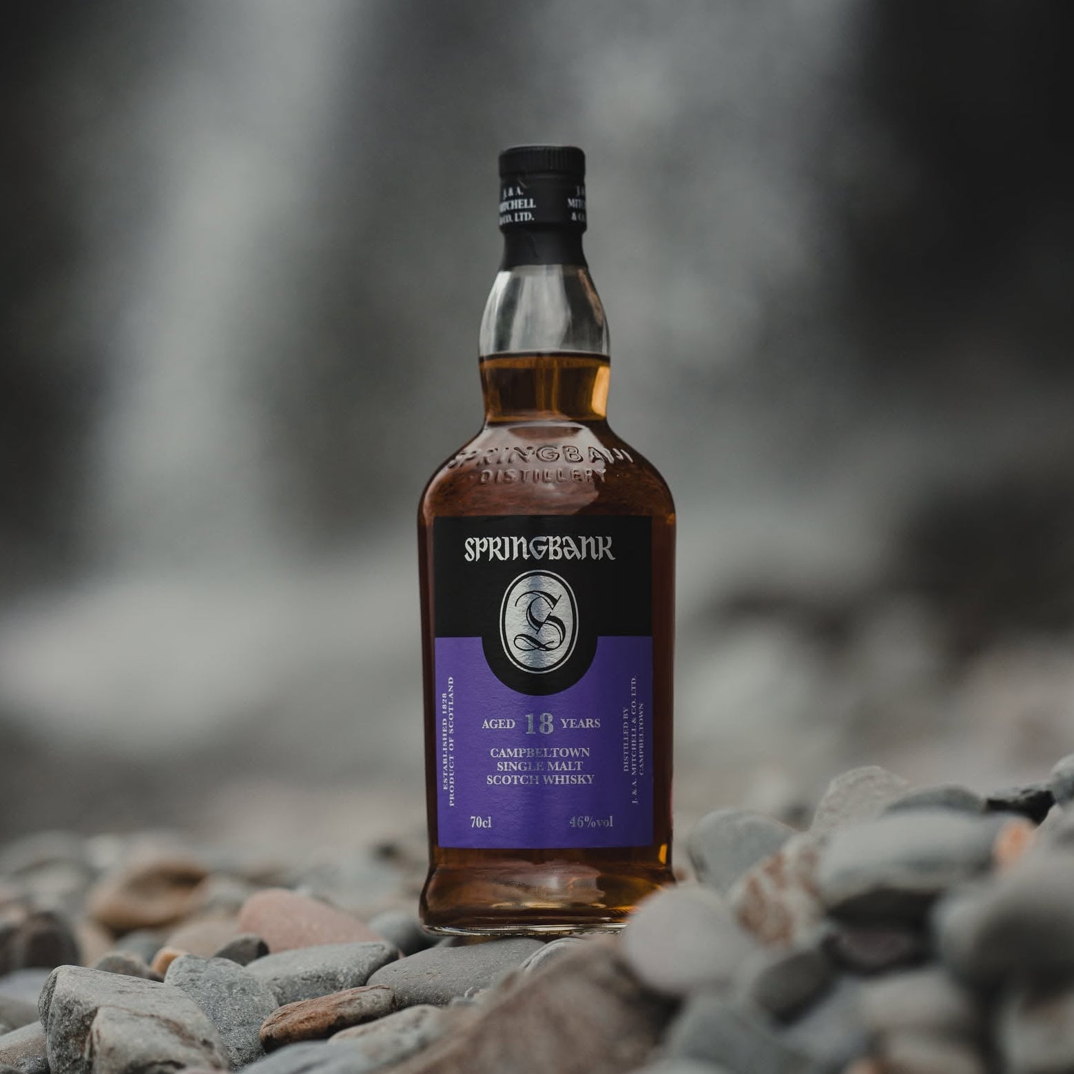 Load image into Gallery viewer, A bottle of Springbank 18 Year Old single malt Scotch whisky stands on a bed of smooth river stones with a blurred waterfall in the background.