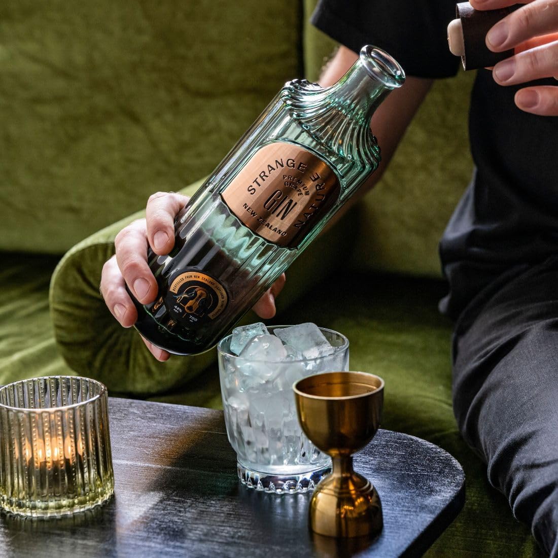 Load image into Gallery viewer, Person holding a bottle of Strange Gin with a glass of ice and a gold cup on a table.