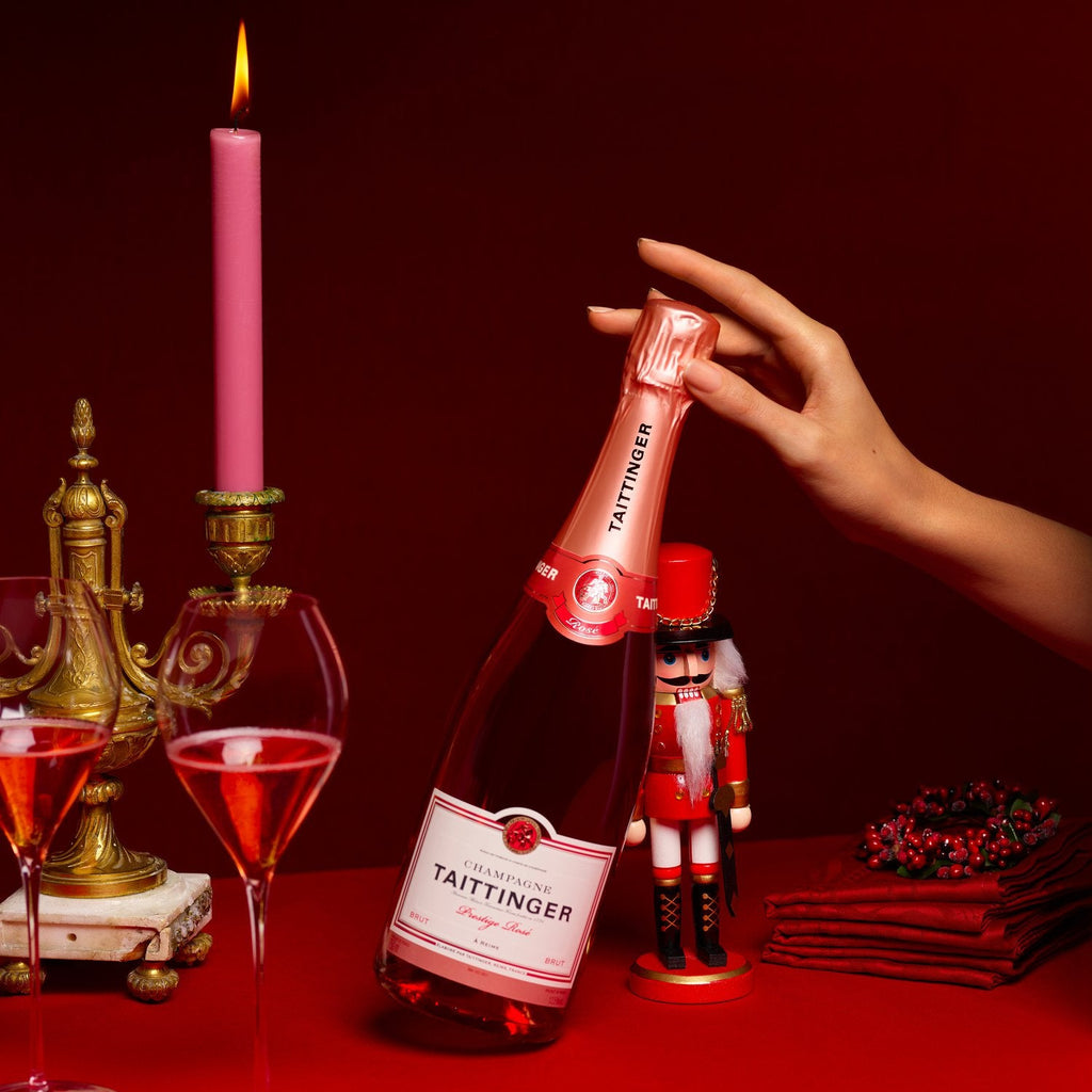 Bottle of Taittinger champagne on a red surface with a hand holding it, candles, and glasses in the background.