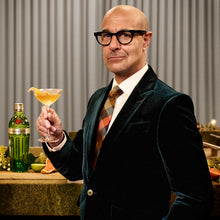 Load image into Gallery viewer, Man in a suit holding a cocktail glass with a bottle and fruit on a bar counter