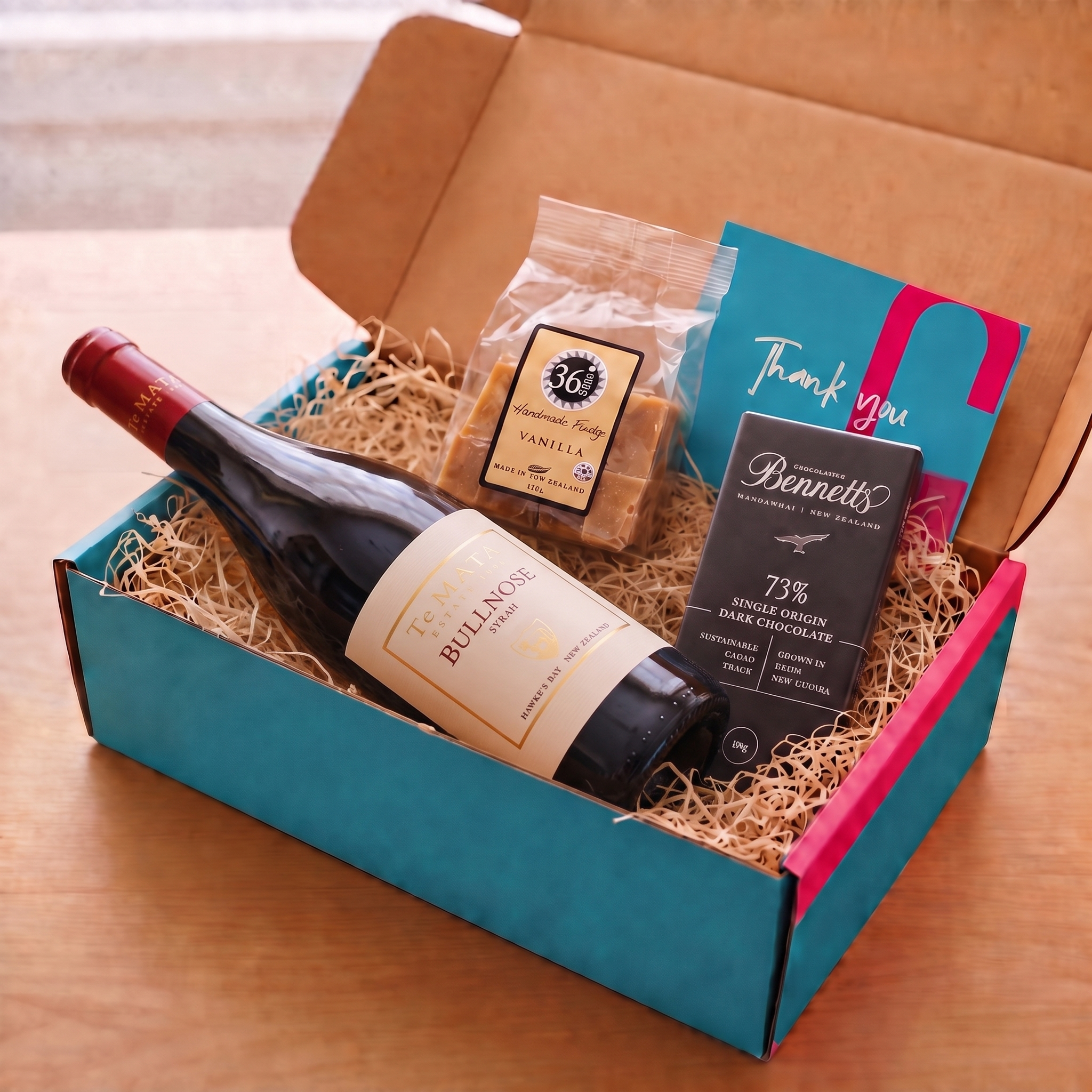 Load image into Gallery viewer, A curated gift box set on a wooden table, featuring a bottle of Te Mata Estate Bullnose Syrah Red Wine, a bag of 36 Degrees Vanilla fudge, a Bennett's 73% Single Origin New Zealand Dark Chocolate bar, and a teal 'Thank you' card inside a brown cardboard box lined with wood-wool filler.