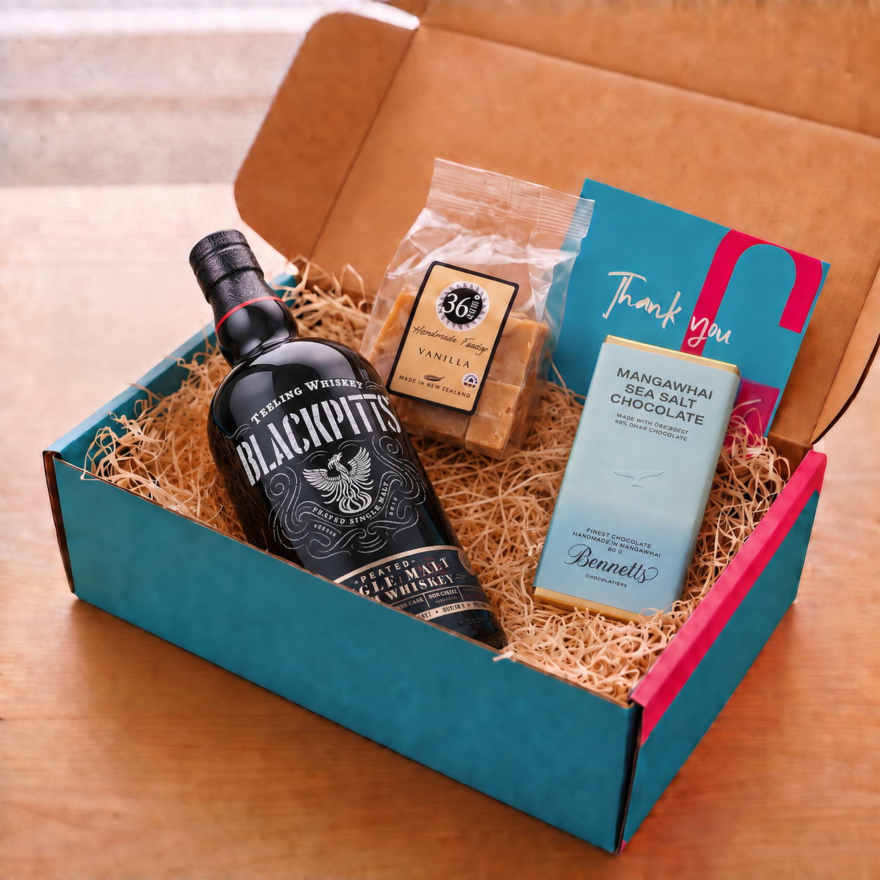 Load image into Gallery viewer, A curated gift box set on a wooden table, featuring a bottle of Teeling Blackpitts Peated Single Malt Irish Whiskey, a bag of 36 Degrees Vanilla fudger, a bar of Bennett's Mangawhai Sea Salt Chocolate, and a blue and pink Thank You card, all arranged in a teal and pink cardboard COVE 27 branded box filled with wood-wool.