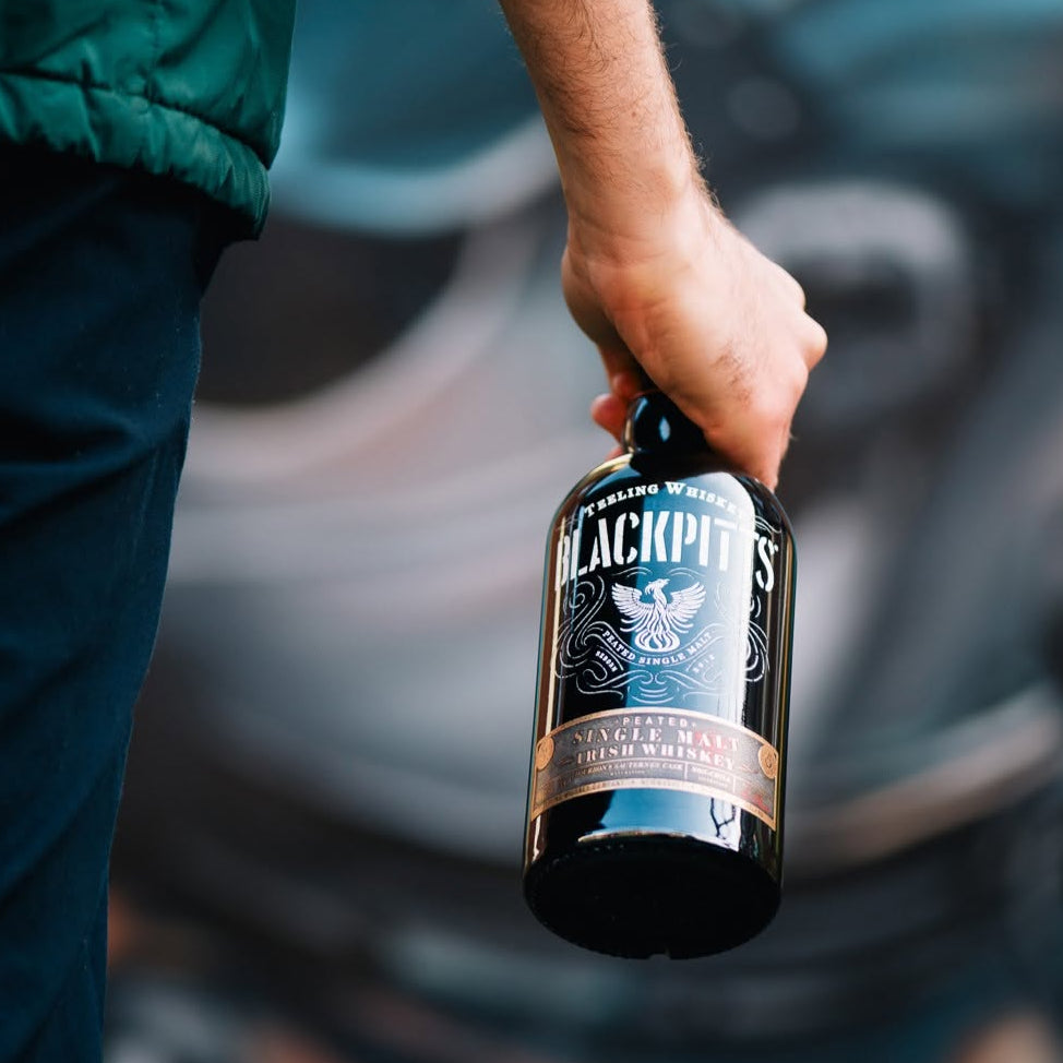 Load image into Gallery viewer, A person holding a bottle of Teeling Blackpitts Peated Single Malt Irish Whiskey against a blurred dark background.