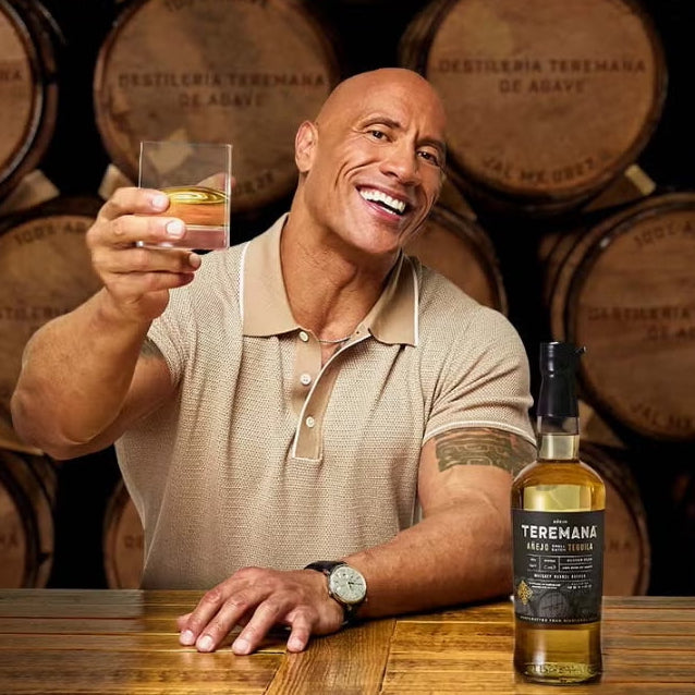 Load image into Gallery viewer, Dwayne 'The Rock' Johnson holding a glass of tequila in front of a bottle of Teremana anejo tequila with wooden barrels in the background