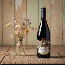 Load image into Gallery viewer, Terra-Sancta-Pinot-Noir-lifestyle wine bottle with decorative label on a wooden surface next to a vase of flowers.
