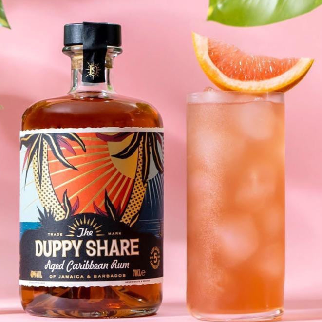 Load image into Gallery viewer, Bottle of 'The Duppy Share' rum, a bottle of pink grapefruit soda, and a glass of pink cocktail with a slice of fruit on a pink background.