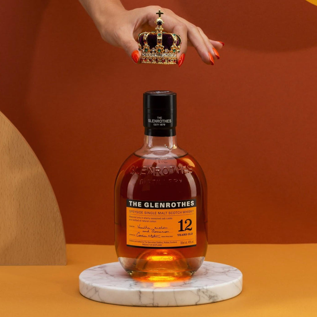 Bottle of Glenrothes whisky on a marble coaster with a hand holding a crown above it against an orange background