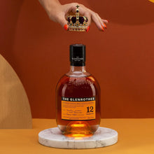 Load image into Gallery viewer, Bottle of Glenrothes whisky on a marble coaster with a hand holding a crown above it against an orange background