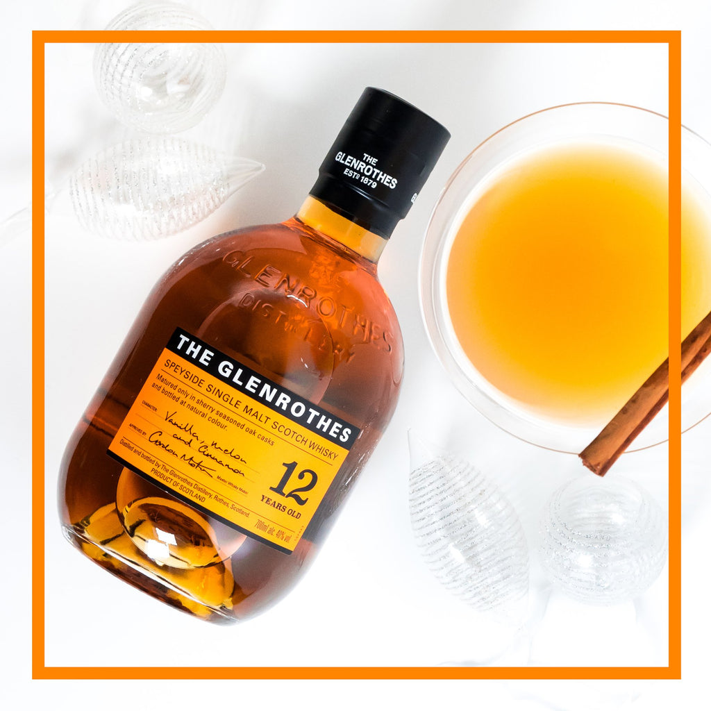 Bottle of The Glenrothes whisky with a glass of orange liquid and cinnamon stick on a white background