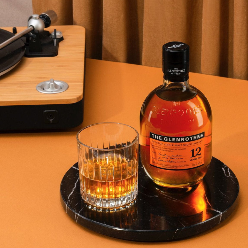 Bottle of Glenrothes whiskey and glass on a turntable with vinyl record, against a brown curtain background.