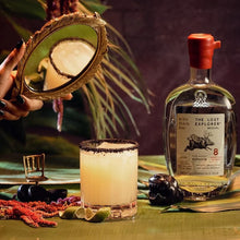 Load image into Gallery viewer, A bottle of mezcal and a margarita cocktail on an inviting green background.