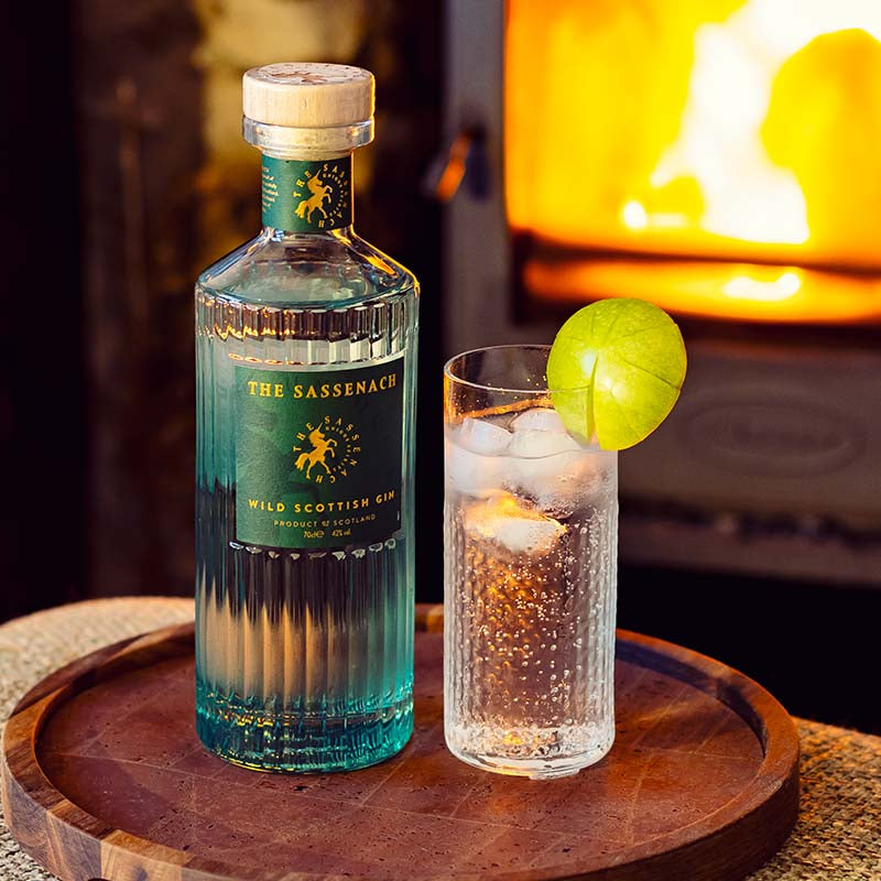 Bottle of The Sassenach gin with a cocktail on a wooden tray in front of a lit fireplace.