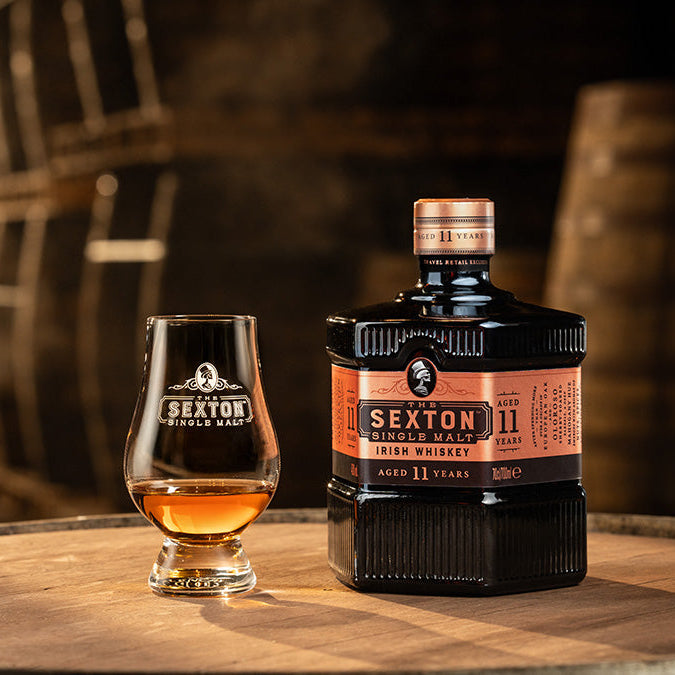 Load image into Gallery viewer, The Sexton 11 Year Old Single Malt Irish Whiskey bottle and glass on a wooden surface with barrels in the background