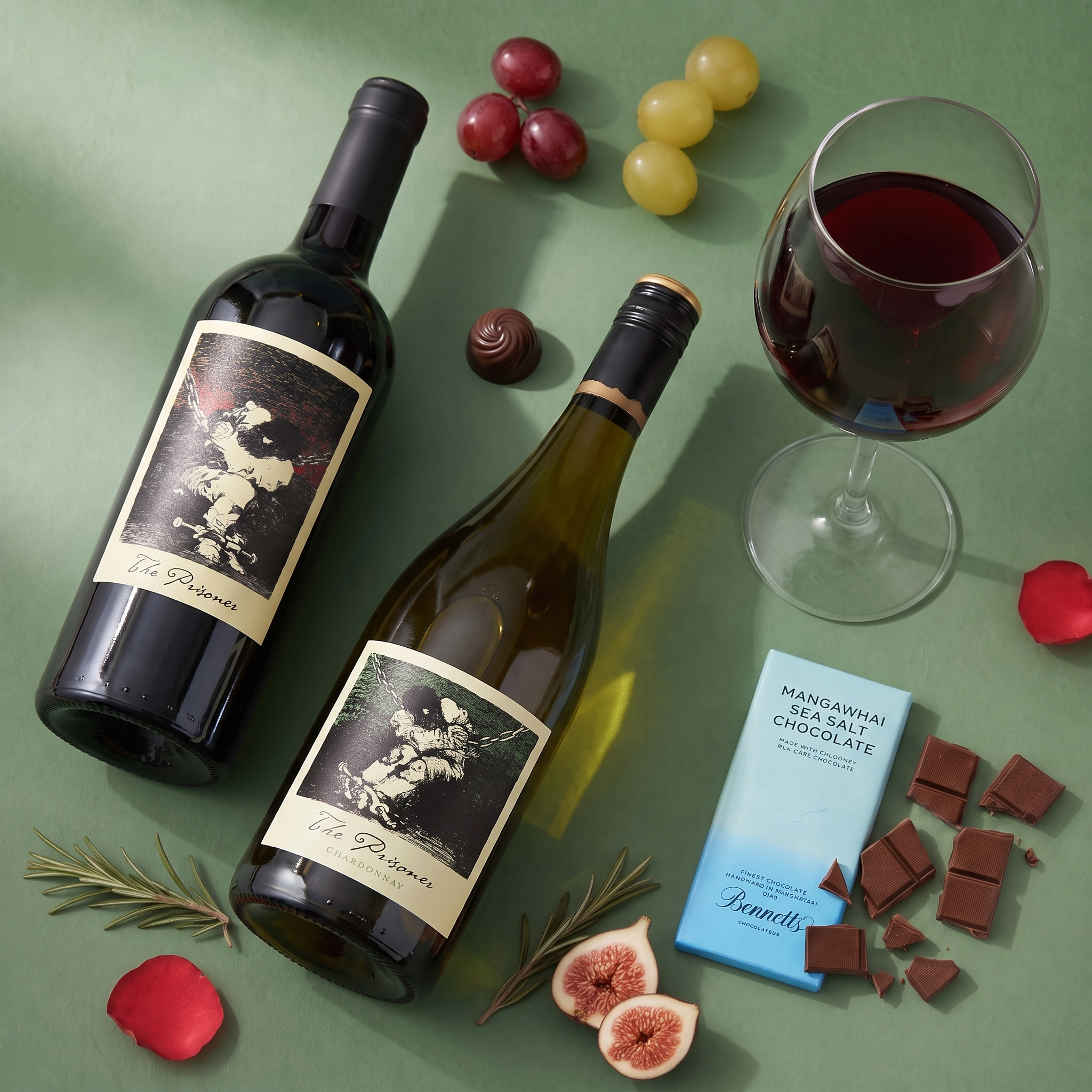 Load image into Gallery viewer, Two wine bottles, The Prisoner Californian Red Blend and Chardonnay white wine arranged on a green surface with a glass of red wine, grapes, figs, chocolate pieces, rose petals, and a blue bar of Benedetto chocolate.