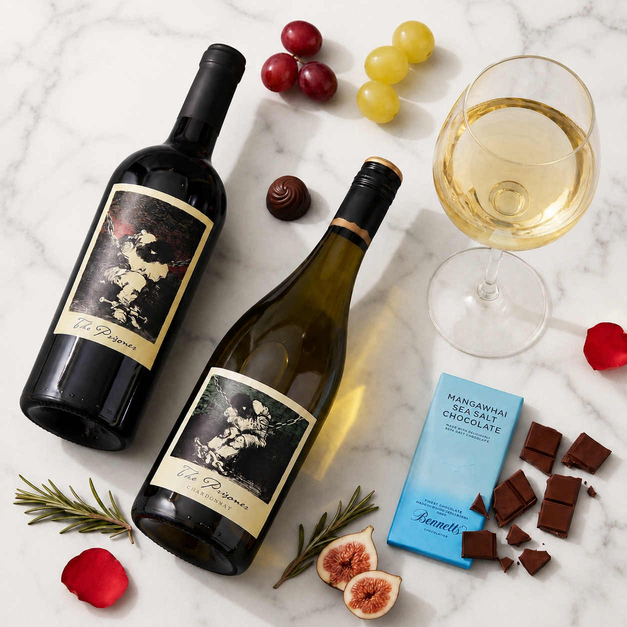 Load image into Gallery viewer, Two bottles of wine, The Prisoner Californian Red Blend and Chardonnay white wine and a filled wine glass arranged on a white marble surface with grapes, chocolates, figs, rosemary, and a blue Bennett chocolate bar.