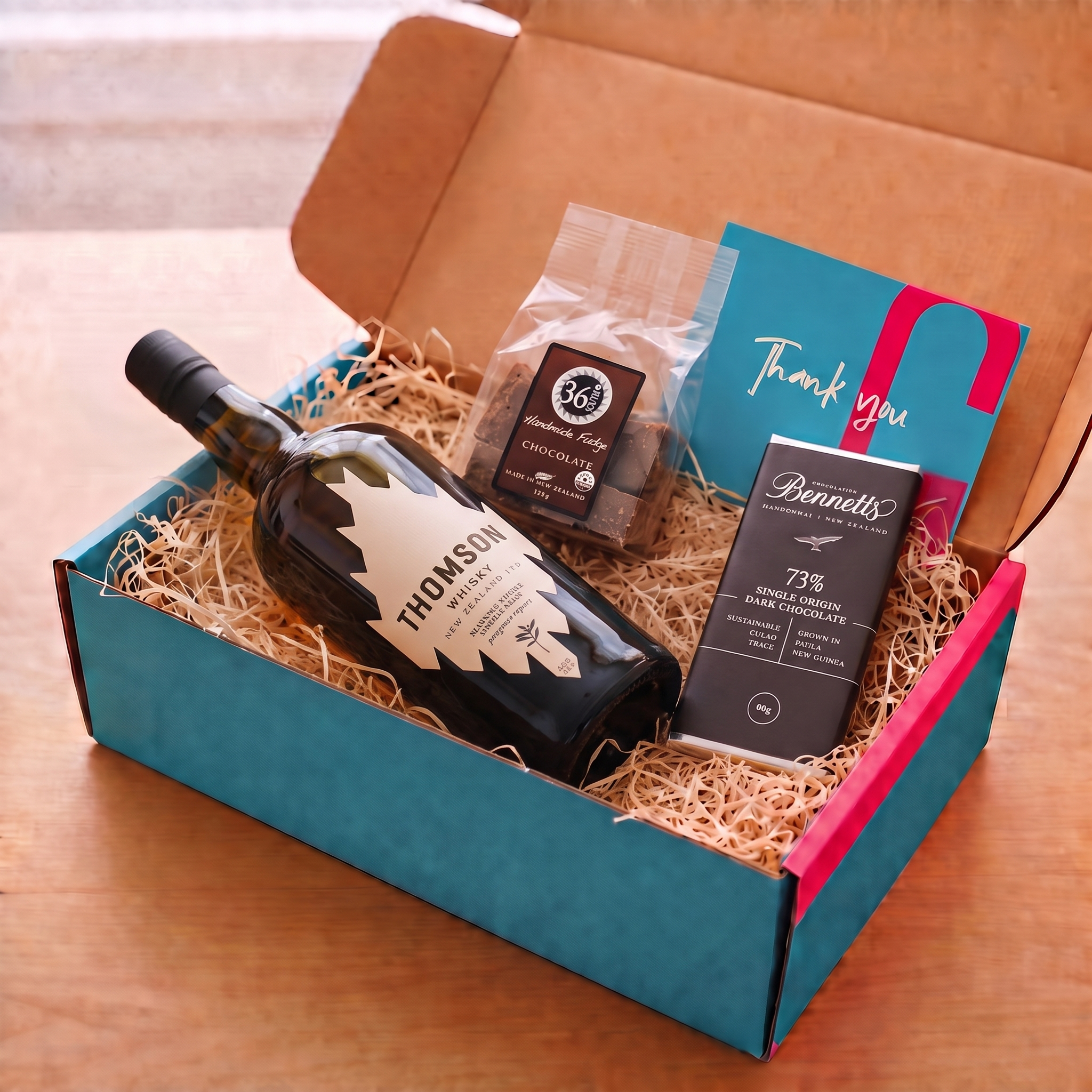 Load image into Gallery viewer, An open gift box on a wooden table containing a bottle of Thomson Manuka Smoke Whisky, a bag of 36 Degrees chocolate fudge, a bar of Bennett's 73% dark chocolate, and a teal thank you card.