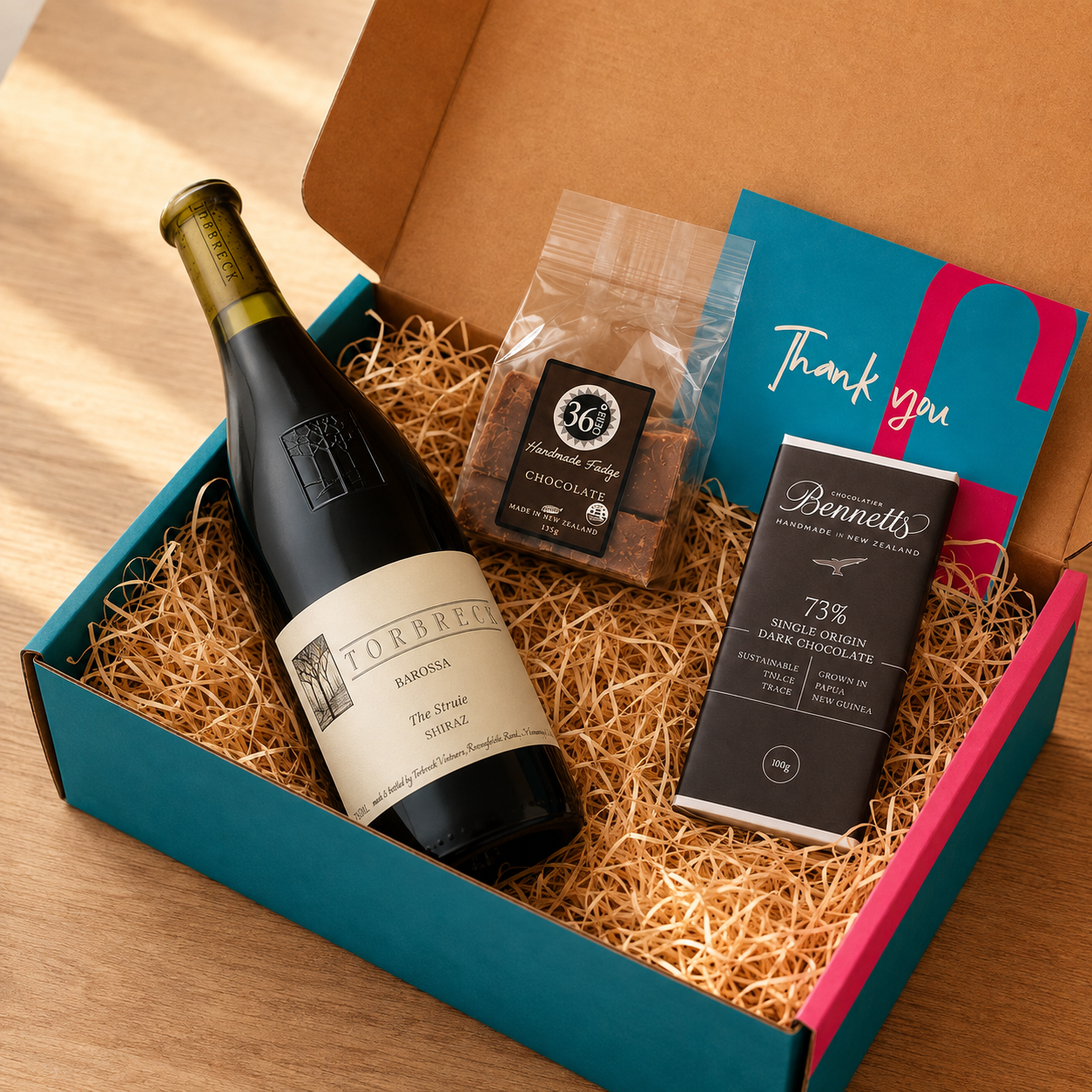 Load image into Gallery viewer, A curated gift box set on a wooden table, containing a bottle of Torbreck 'The Struie' Barossa Shiraz red wine, a bag of 36 Degrees chocolate fudge, a bar of Bennetts 73% single origin dark chocolate, and a blue and pink 'Thank you' card, all arranged in brown paper shred inside a teal and pink box.