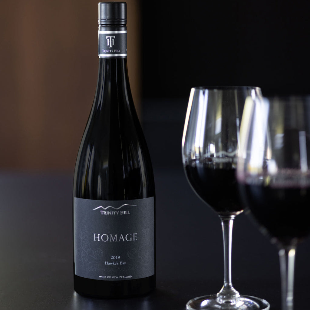Bottle of A bottle of Trinity Hill 'Homage' Syrah wine with a black label featuring the Trinity Hill logo. wine with two glasses on a dark surface