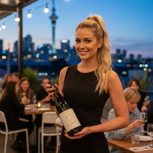 Load image into Gallery viewer, A waitress holding a bottle of Trinity Hill 125 Chardonnay wine in a restaurant with the Auckland skyline in the background.