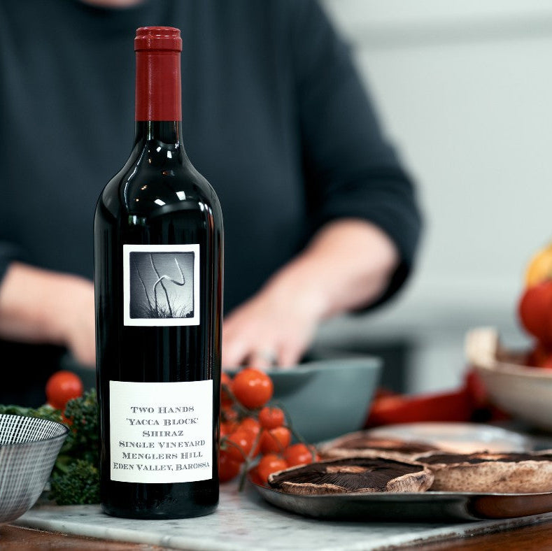 Load image into Gallery viewer, Bottle of Two Hands Yacca Block Shiraz red wine with a blurred background of a person and food items.