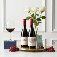 Load image into Gallery viewer, Two bottles of Two Paddocks Pinot Noir red wine on a tray with a glass, Bennetts Milk Chocolate bar, grapes, and flowers against a white wall.