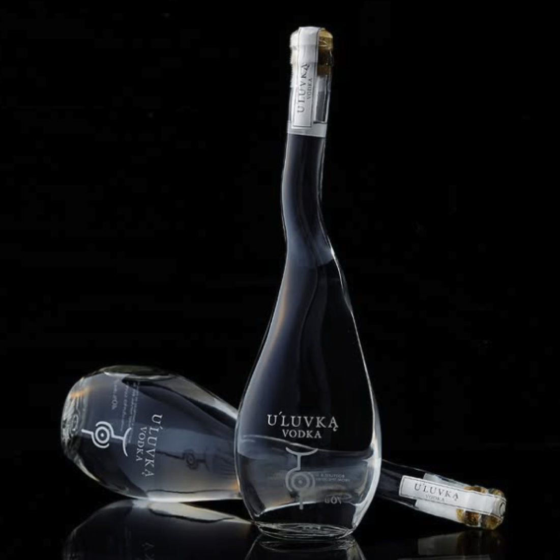 Load image into Gallery viewer, Three clear glass bottles of U'Luvka Vodka arranged on a glossy black reflective surface against a black background, with one standing upright and two lying on their sides.