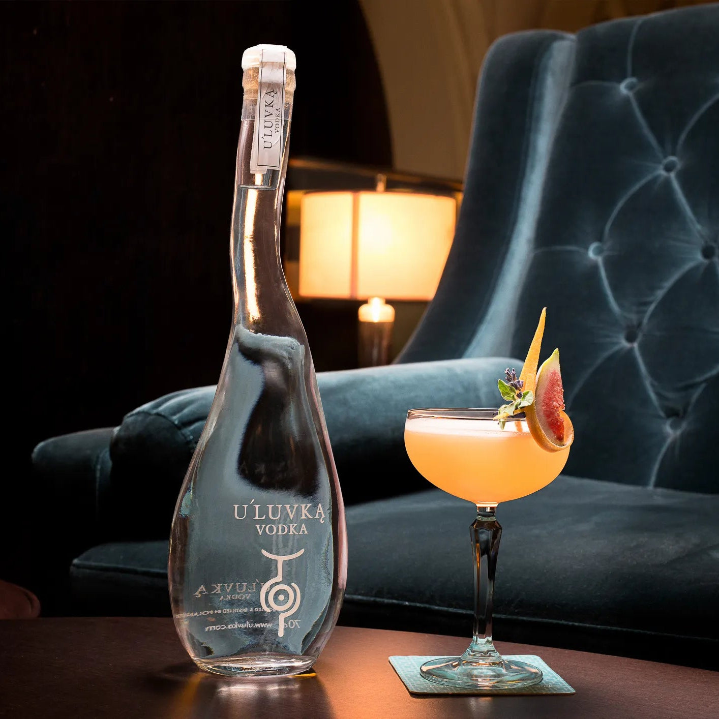 Load image into Gallery viewer, A clear glass bottle of U'Luvka Vodka stands on a table beside a filled orange cocktail in a coupe glass, with a dark tufted velvet armchair and warm lamp in the background.