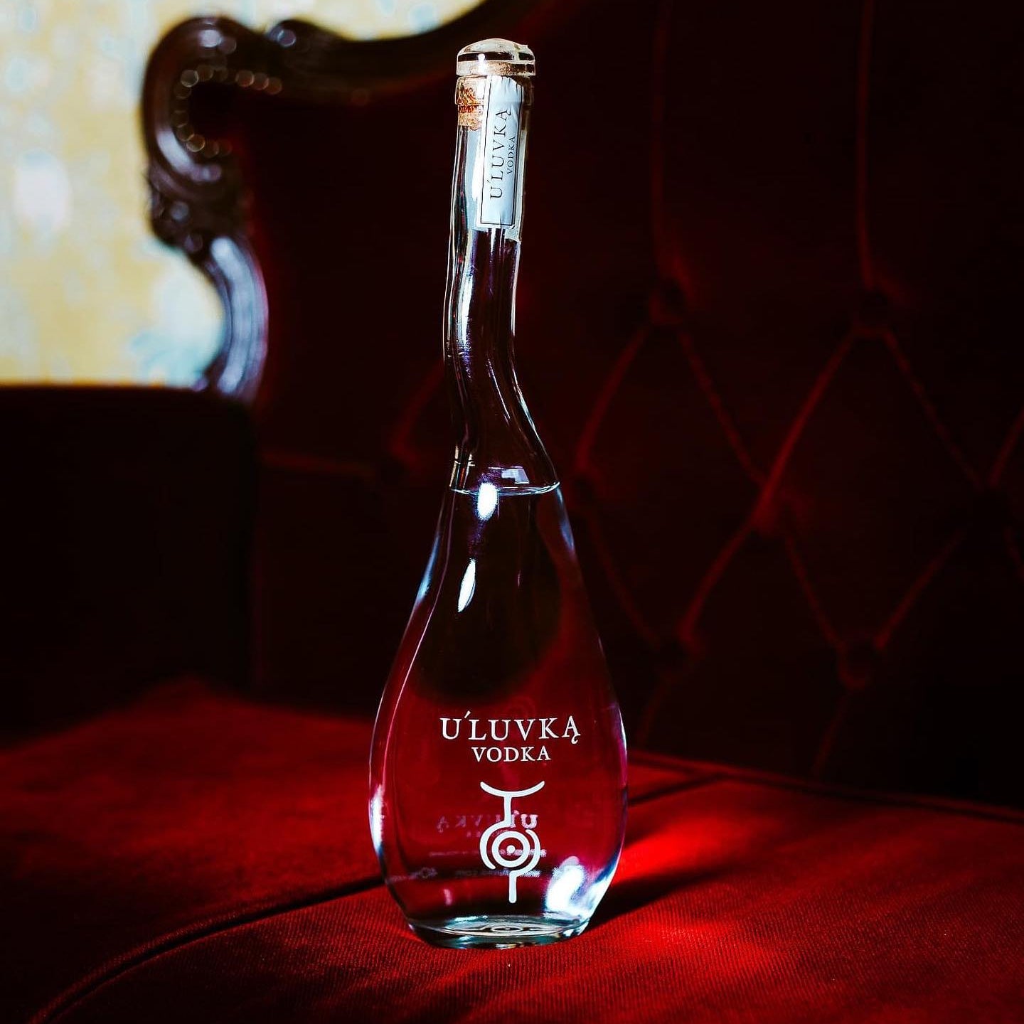 Load image into Gallery viewer, A bottle of U'Luvka Vodka, standing on a red velvet surface in front of a dark red tufted chair.