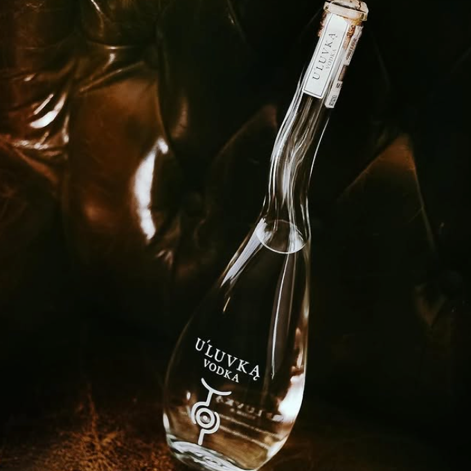 Load image into Gallery viewer, A clear glass bottle of U'Luvka Vodka with a silver foil cap, set against a dark, moody background with warm brown highlights.