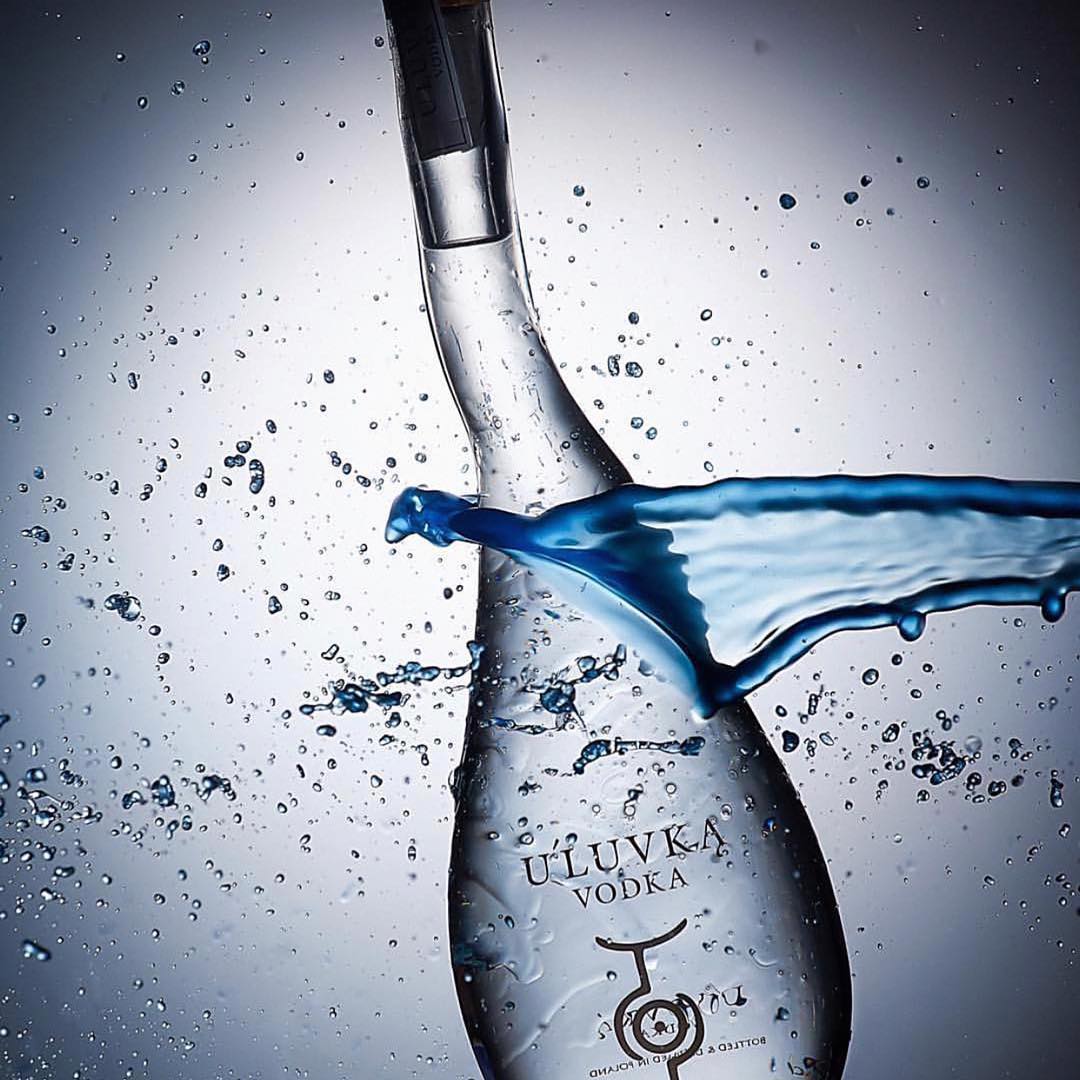 Load image into Gallery viewer, A bottle of U'Luvka Vodka with blue liquid splashing across it, photographed against a cool grey background with water droplets and a soft glow.