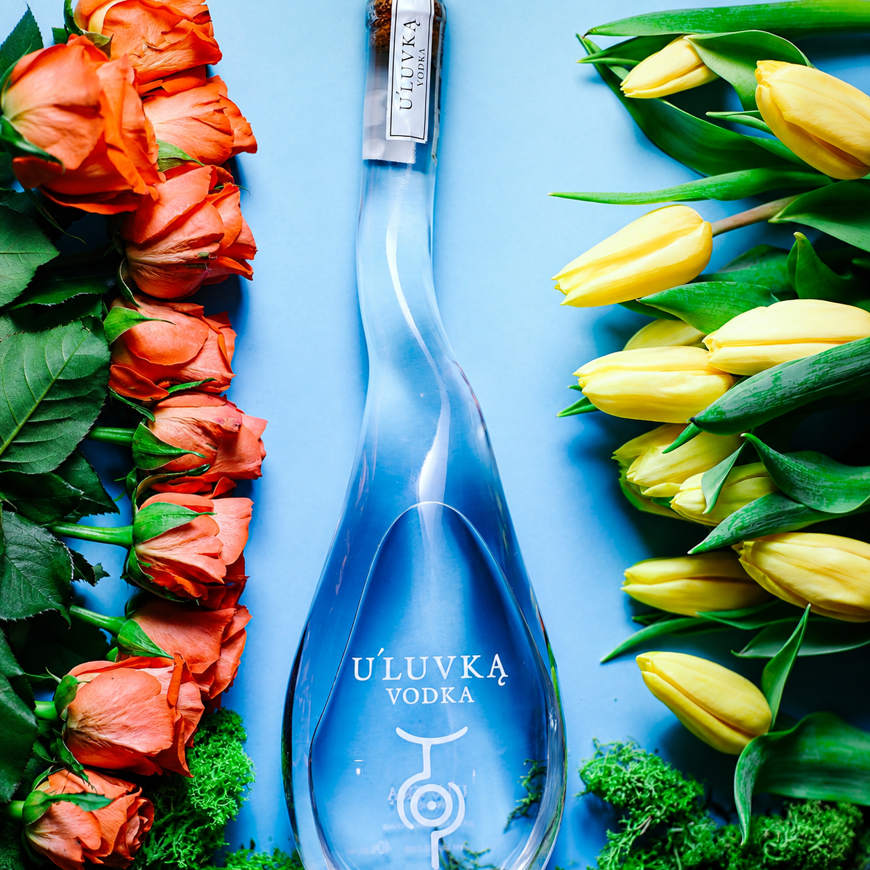 Load image into Gallery viewer, A bottle of U'Luvka Vodka is centred on a light blue background, framed by orange roses on the left and yellow tulips with green leaves on the right.