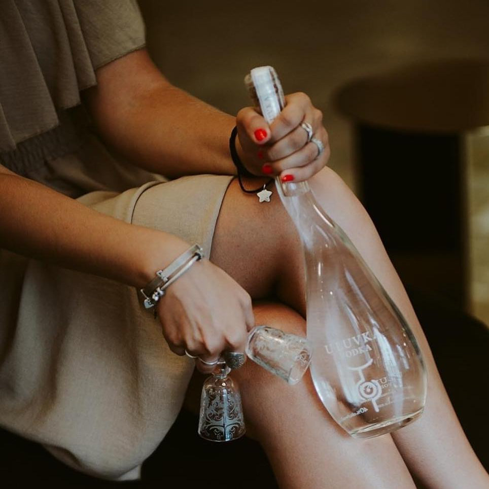 Load image into Gallery viewer, A woman in a beige dress and black pointed-toe heels sits on a dark sofa, holding a bottle of U'Luvka Vodka in a dimly lit indoor setting.