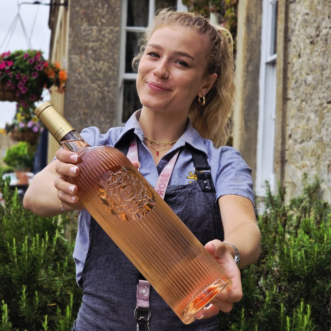 Load image into Gallery viewer, Woman holding a large bottle of rose outdoors with a building and flowers in the background