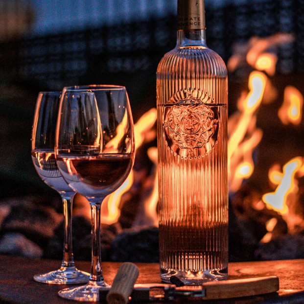 Load image into Gallery viewer, A bottle of Ultimate Provence rosé wine with two filled wine glasses, a corkscrew, and a matchbox set against a warm fire at dusk.