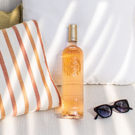 Load image into Gallery viewer, A bottle of Ultimate Provence rosé wine stands on a white textured sofa with a large white pillow behind it, a striped throw pillow to the left, and sunglasses in the foreground under dappled sunlight.