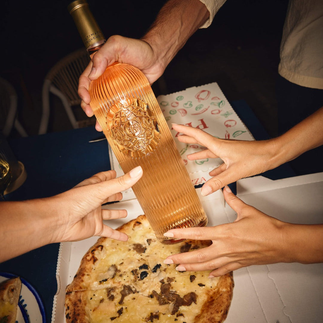 Load image into Gallery viewer, A group of people's hands and arms are visible as they pour a bottle of Ultimate Provence rosé wine into a box of pizza at a dining table.