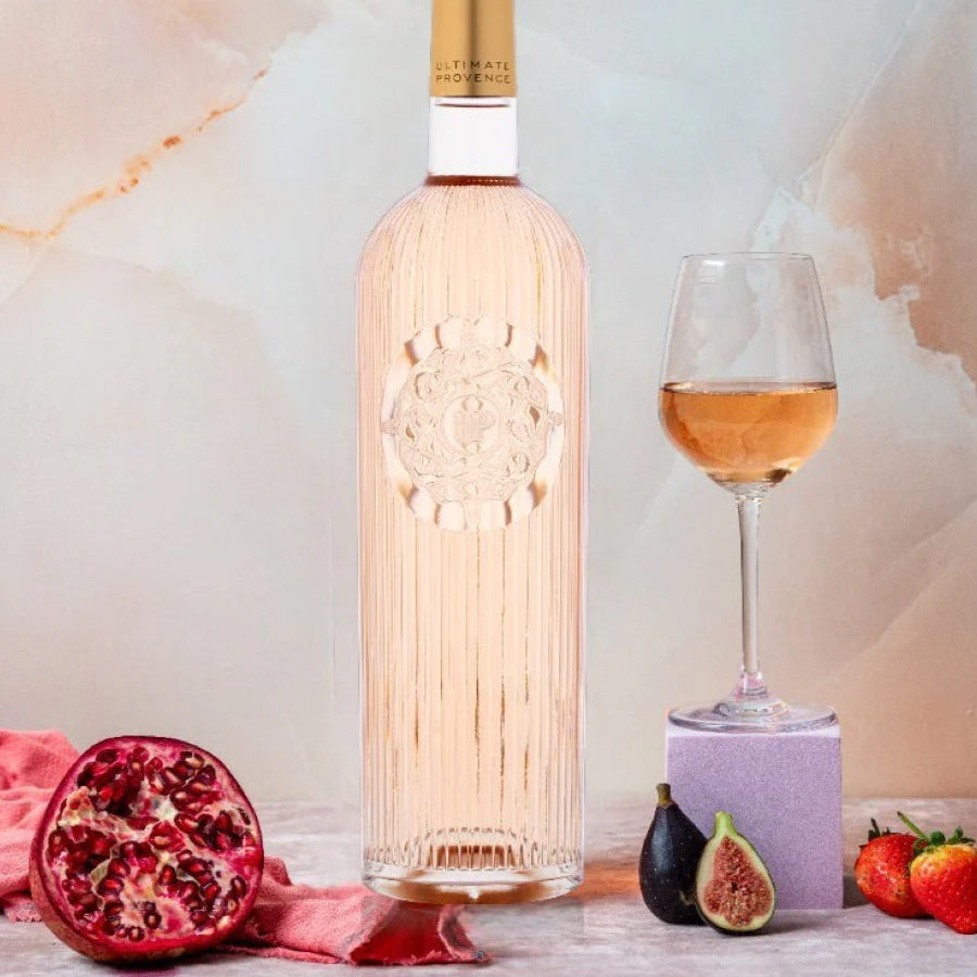 Load image into Gallery viewer, A bottle of Ultimate Provence rosé wine with a gold cap and a glass of rose gold wine, styled with fresh fruits including a pomegranate, fig, and strawberries on a marble surface.
