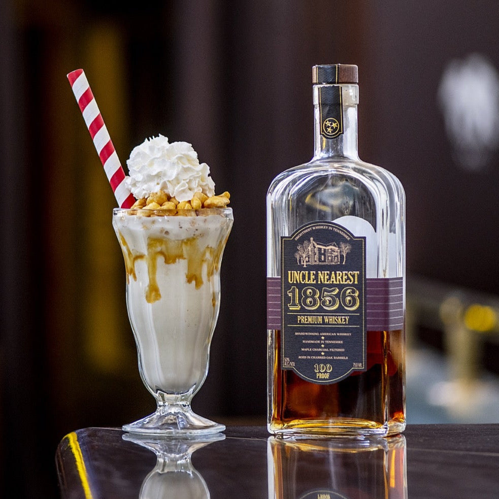 Load image into Gallery viewer, Whiskey bottle with a milkshake and straw on a bar counter.
