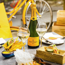 Load image into Gallery viewer, Bottle of Veuve Clicquot with two glasses of champagne on a table with food and a yellow bicycle in the background.