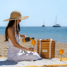 Load image into Gallery viewer, Woman on a beach pouring champagne from a bottle into glasses, with a picnic basket nearby.