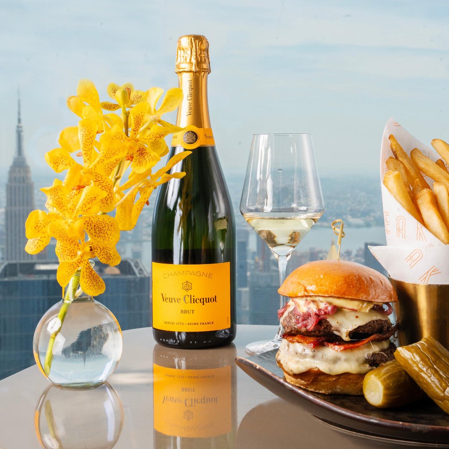 Load image into Gallery viewer, Bottle of Perrier-Jouét champagne, a glass of champagne, a burger, and fries on a table with a cityscape background.
