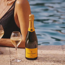 Load image into Gallery viewer, Bottle of Veuve Clicquot champagne with two glasses on a stone ledge by water.