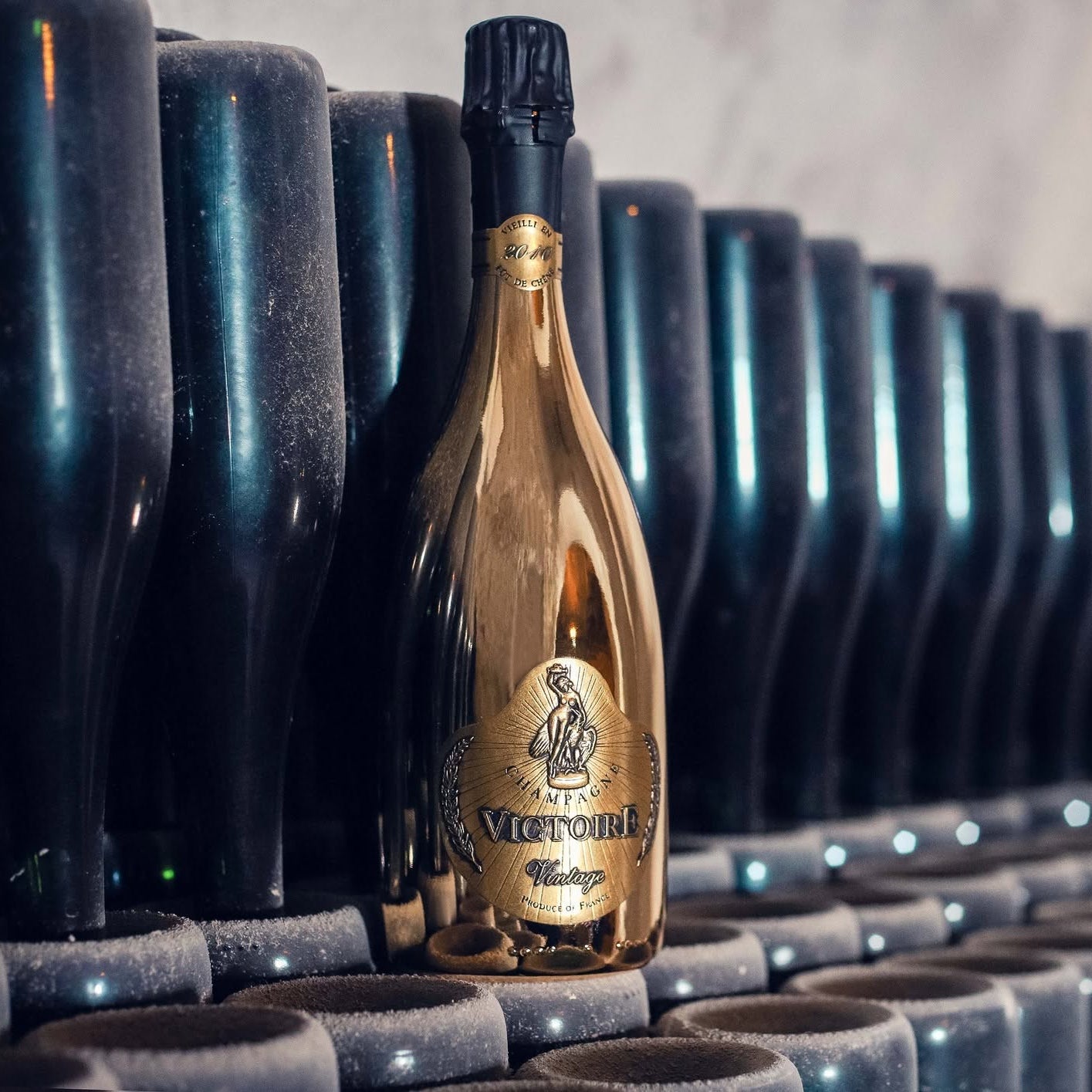 Load image into Gallery viewer, A gold bottle of Victoire Fût de Chêne Prestige Champagne stands upright in a row of dark glass bottles on a stone surface, with shallow depth of field blurring the background.