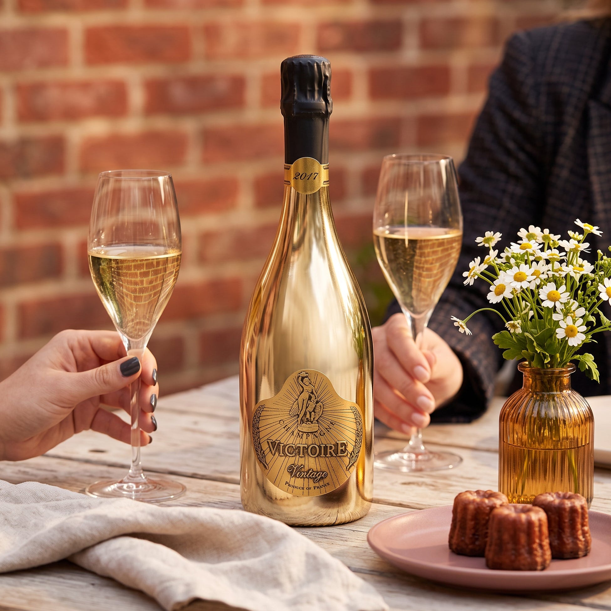 Load image into Gallery viewer, A gold bottle of Victoire Fût de Chêne Prestige Champagne in a shiny gold bottle, styled on a wooden table with two hands holding champagne flutes, a small vase of daisies, and chocolate cakes against a brick wall background.