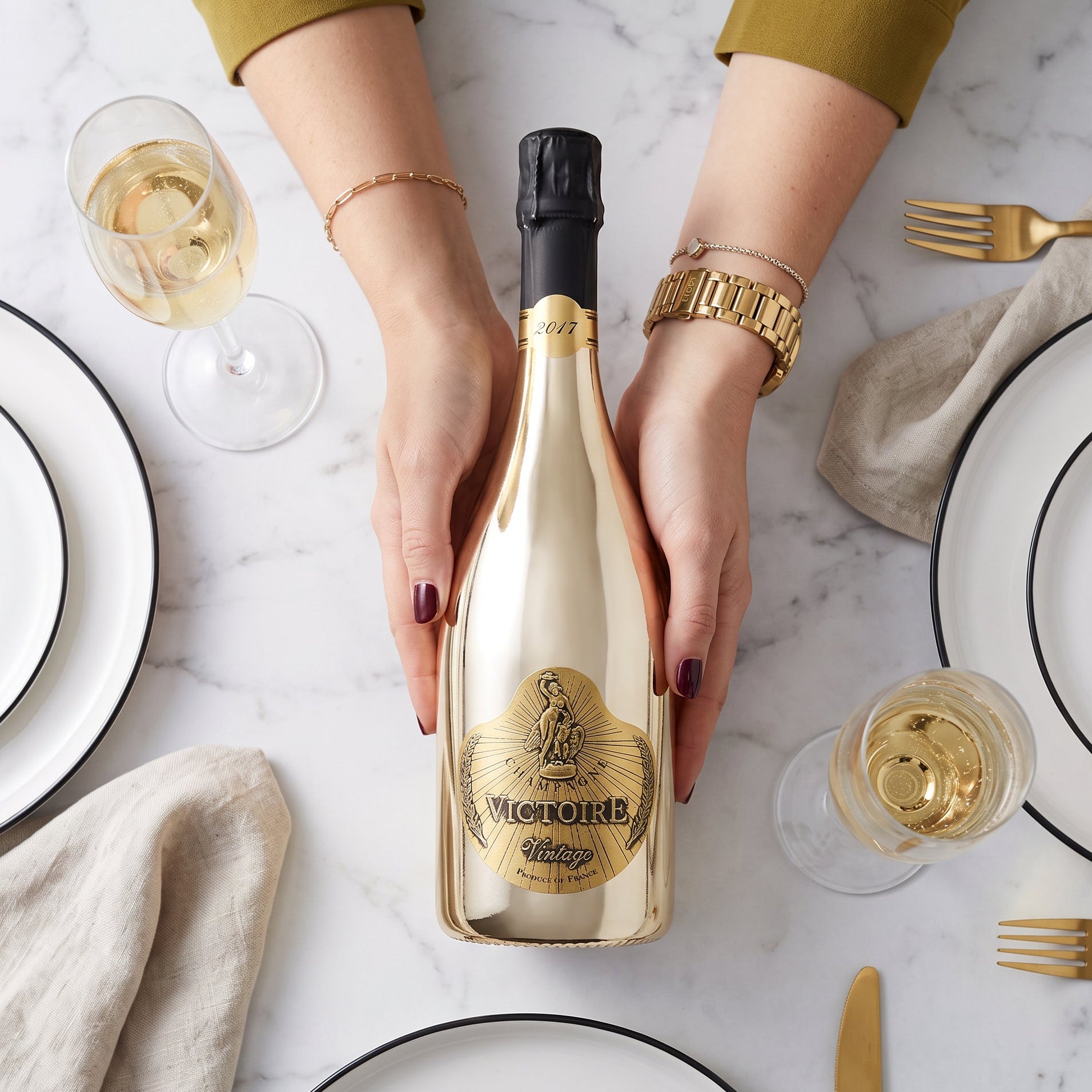 Load image into Gallery viewer, A gold bottle of Victoire Fût de Chêne Prestige Champagne held by two hands over a marble table set with white plates, gold cutlery, and glasses of white wine.