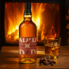 Load image into Gallery viewer, Bottle of Waiheke-Whisky-Dyad-II-Single-Malt-Whisky with a glass of whiskey and ice cubes on a wooden surface with a fireplace in the background.